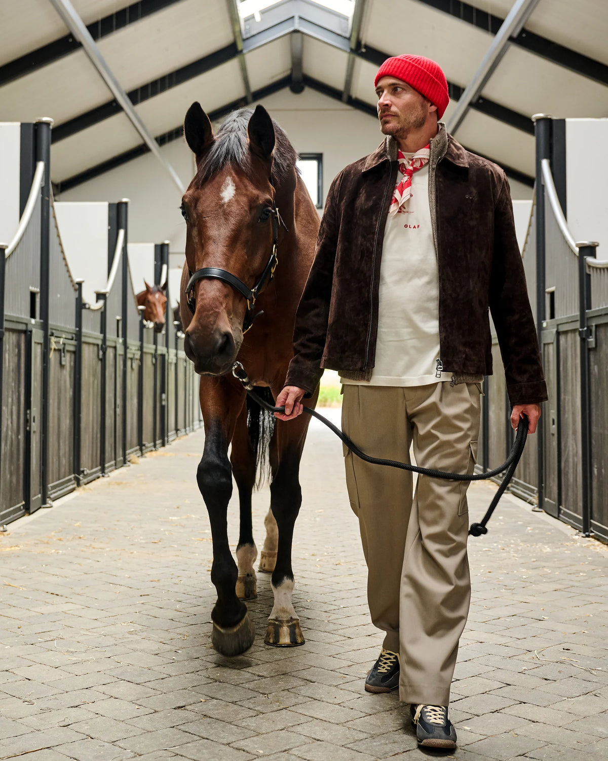 Bruin OLAF suède jasje met lichte lagen, zandkleurige broek en rode muts naast een paard op Begijnhoeve.