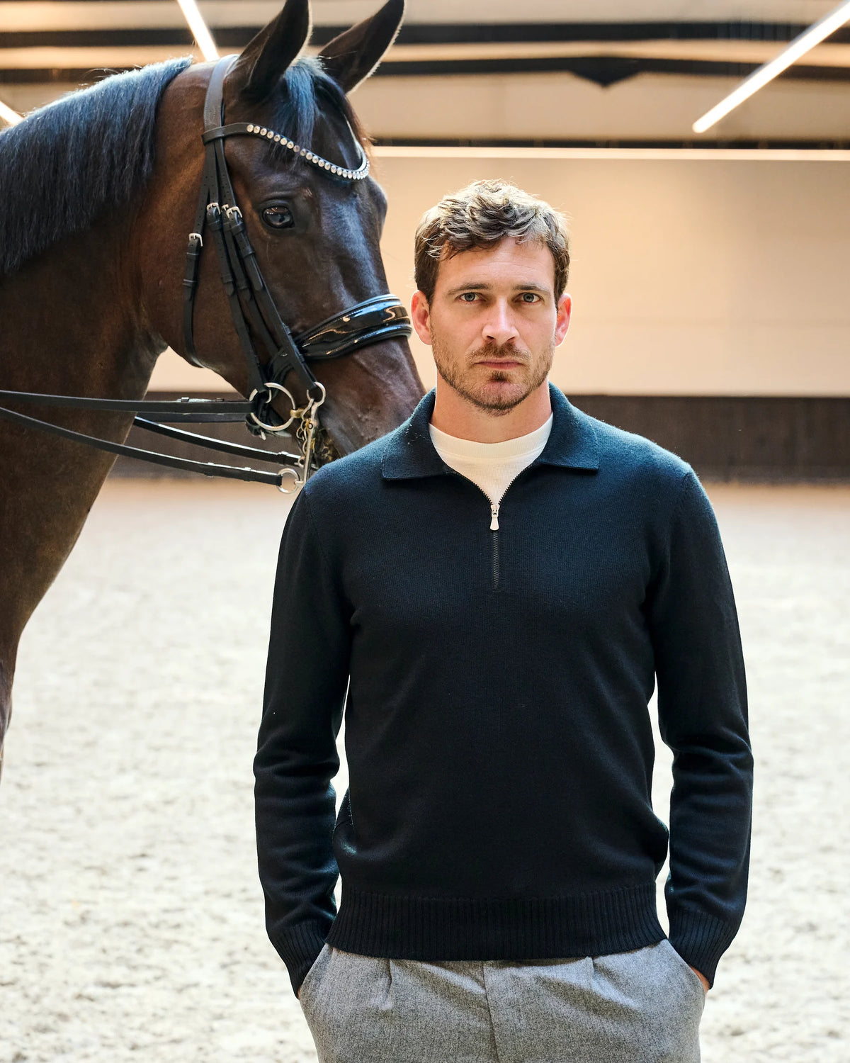 Donkere gebreide zip polo met lichte geruite broek naast een paard in de rijpiste van Begijnhoeve.