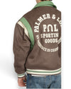 Detail van de PAL-rugborduring, groene schouderpanelen en gestreepte boord op de Ranch Varsity Jacket in Deep Mahogany.