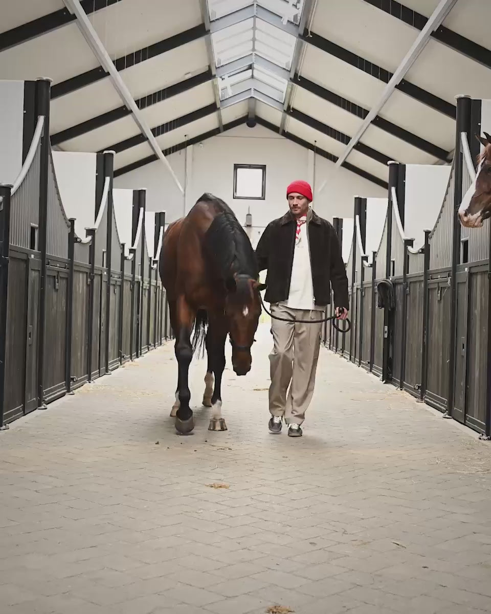 Bruin suède jasje, lichte knitwear, beige broek en rode muts naast een paard in de stallen op Begijnhoeve in Mol-Postel.